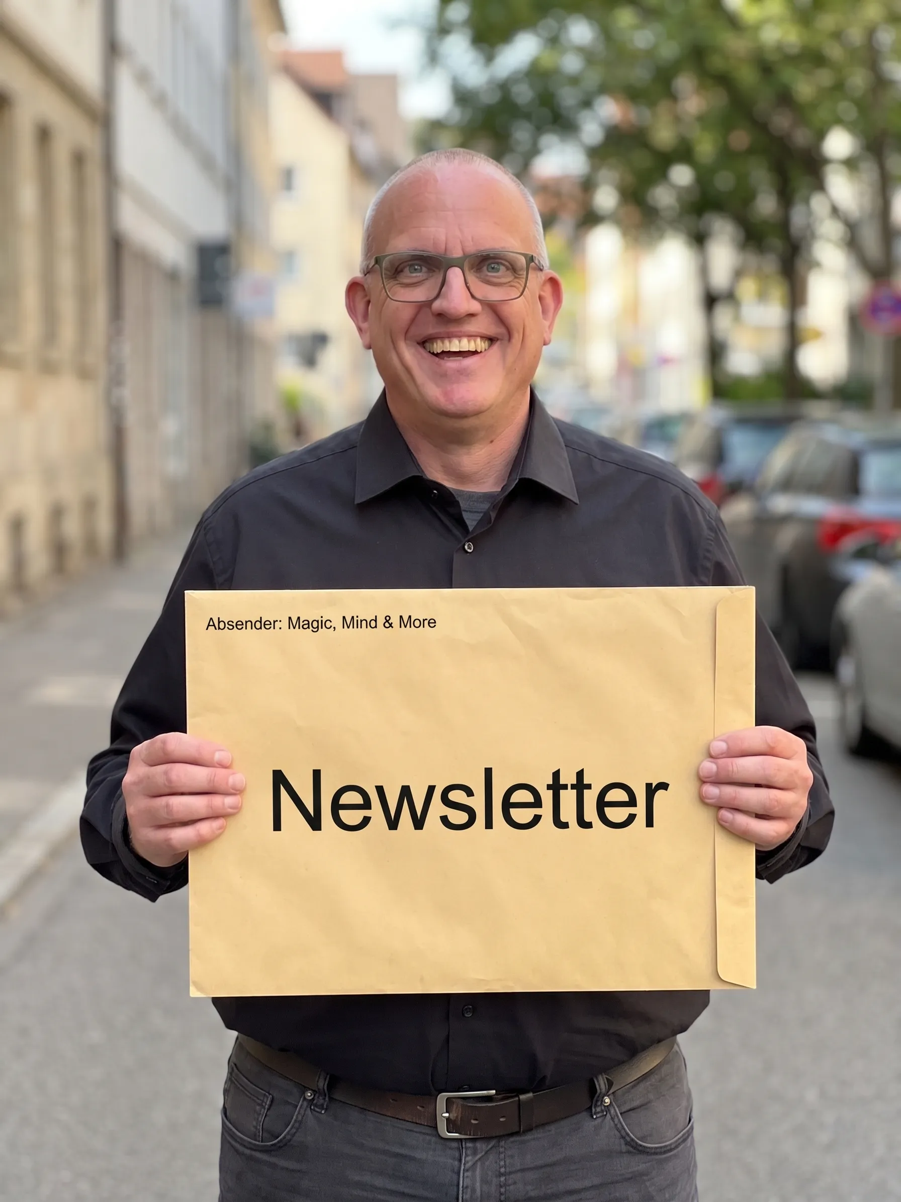 Joachim Jögel laughing in the street, holding a large envelope marked “Newsletter” and “Absender: Magic, Mind &amp; More”