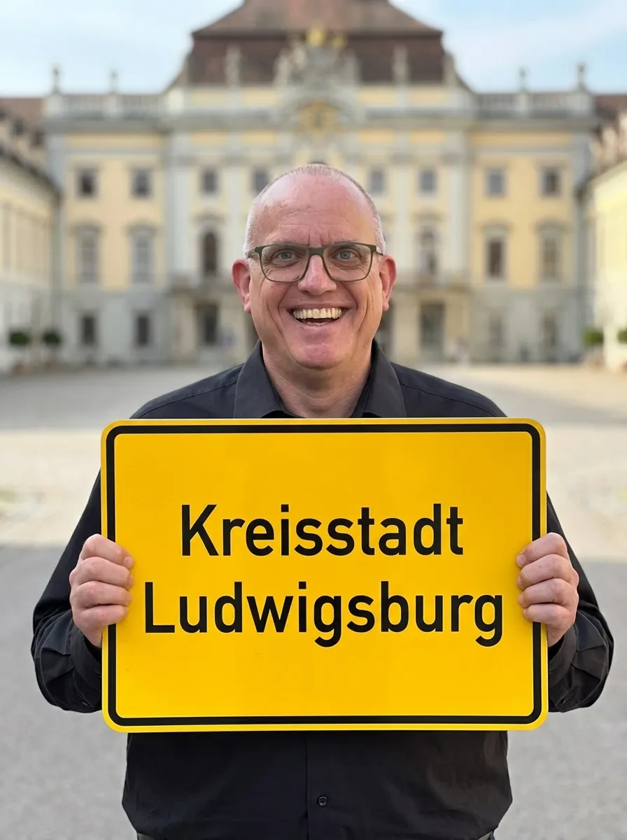 Joachim Jögel lächelnd mit einem Ortsschild „Kreisstadt Ludwigsburg" in den Händen, im Hintergrund das Residenzschloss Ludwigsburg