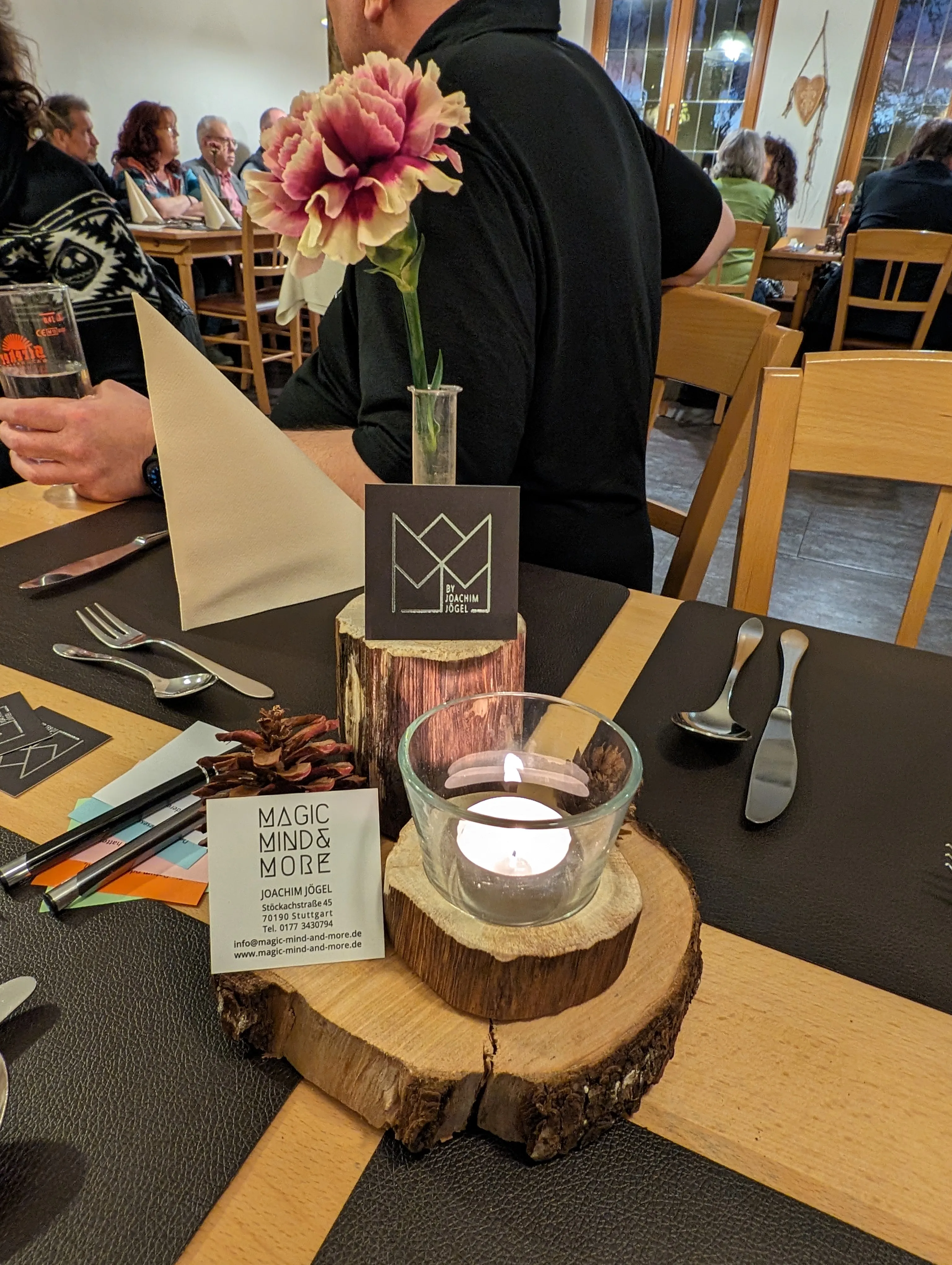 Tischdekoration mit Magic-Mind-&-More-Visitenkarte, Kerze und Blume, im Hintergrund Gäste im Restaurant