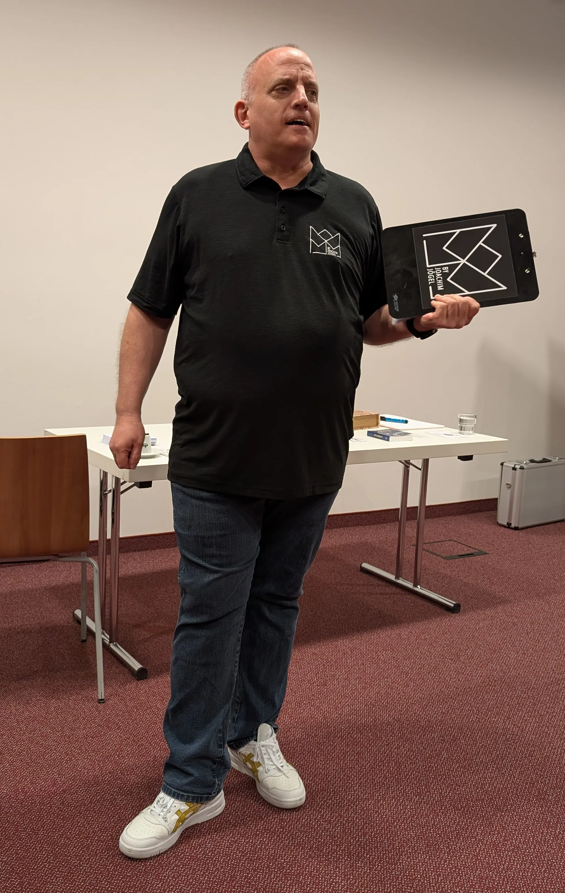 Joachim Jögel giving a talk to a corporate audience, holding a clipboard prop